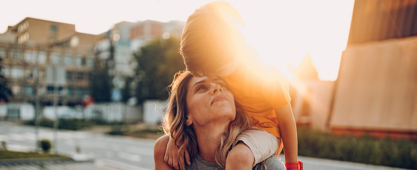 A woman giving a piggyback ride to a child on a sunny city street, both smiling and enjoying each other's company during golden hour.